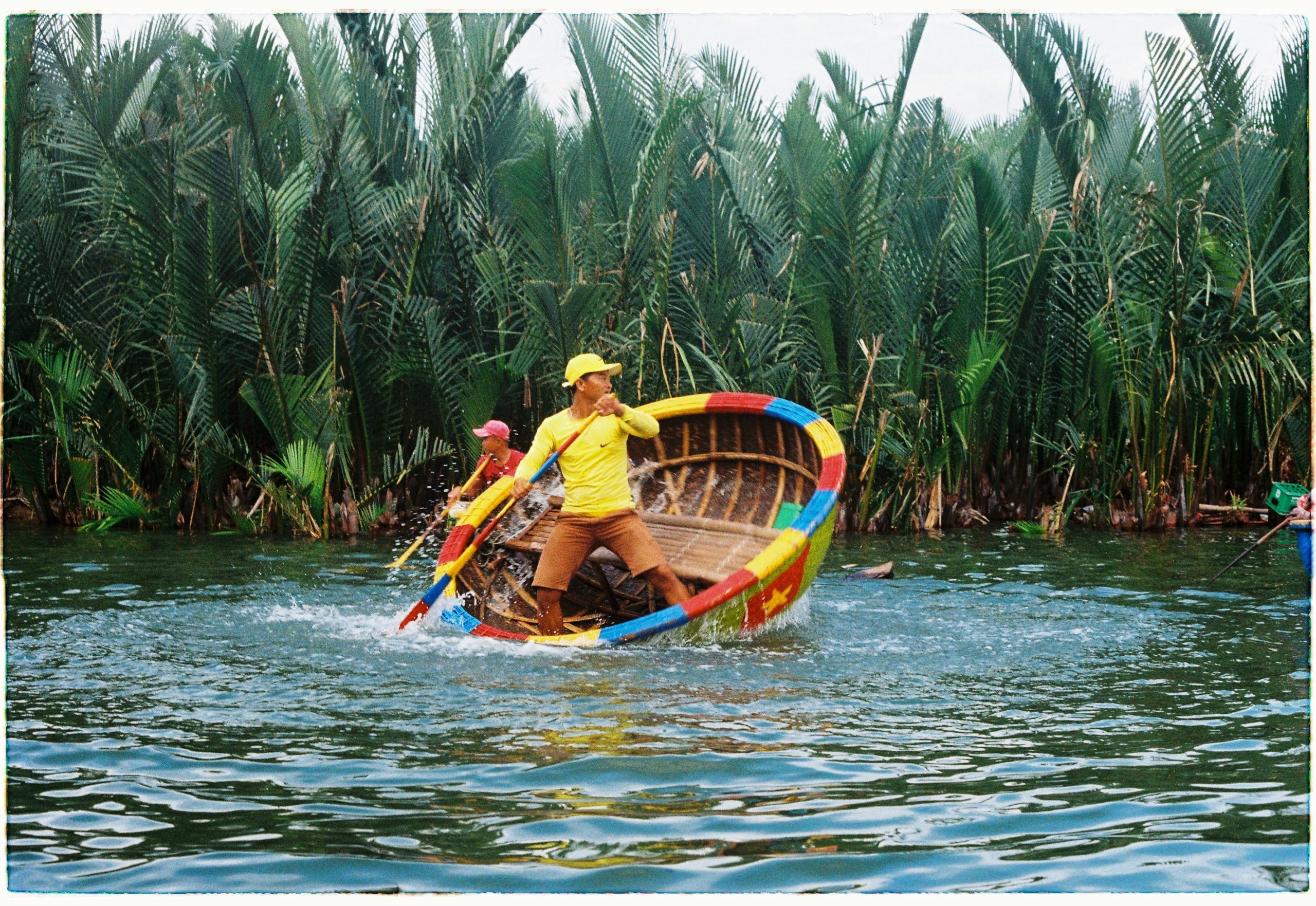 Man navigating a colorful basket boat through lush palm trees in Hoi An, Vietnam, showcasing vibrant culture and scenery.