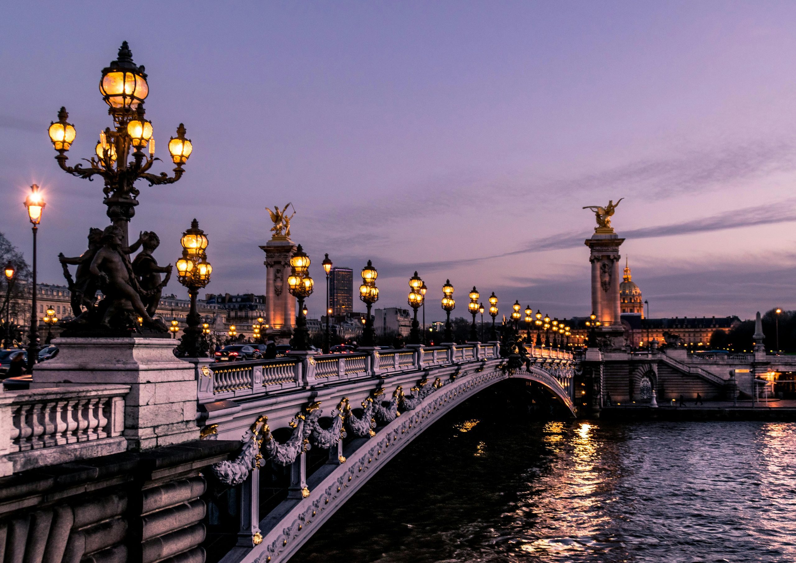 Paris at dusk, showcasing the illuminated Alexandre III Bridge with its ornate sculptures and lampposts over the Seine River.