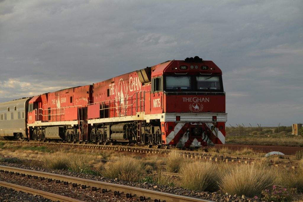 Red passenger train 'The Ghan' traveling on railway track through Australian outback under cloudy sky.
