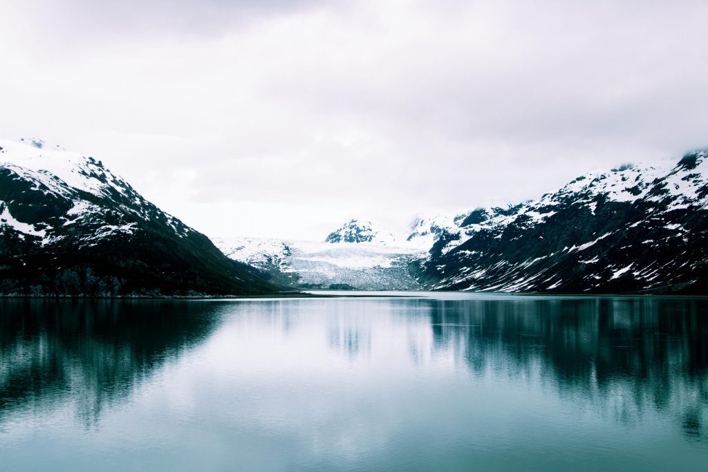 Serene Alaskan glacier reflected in calm icy waters, flanked by snow-capped mountains under a cloudy sky.