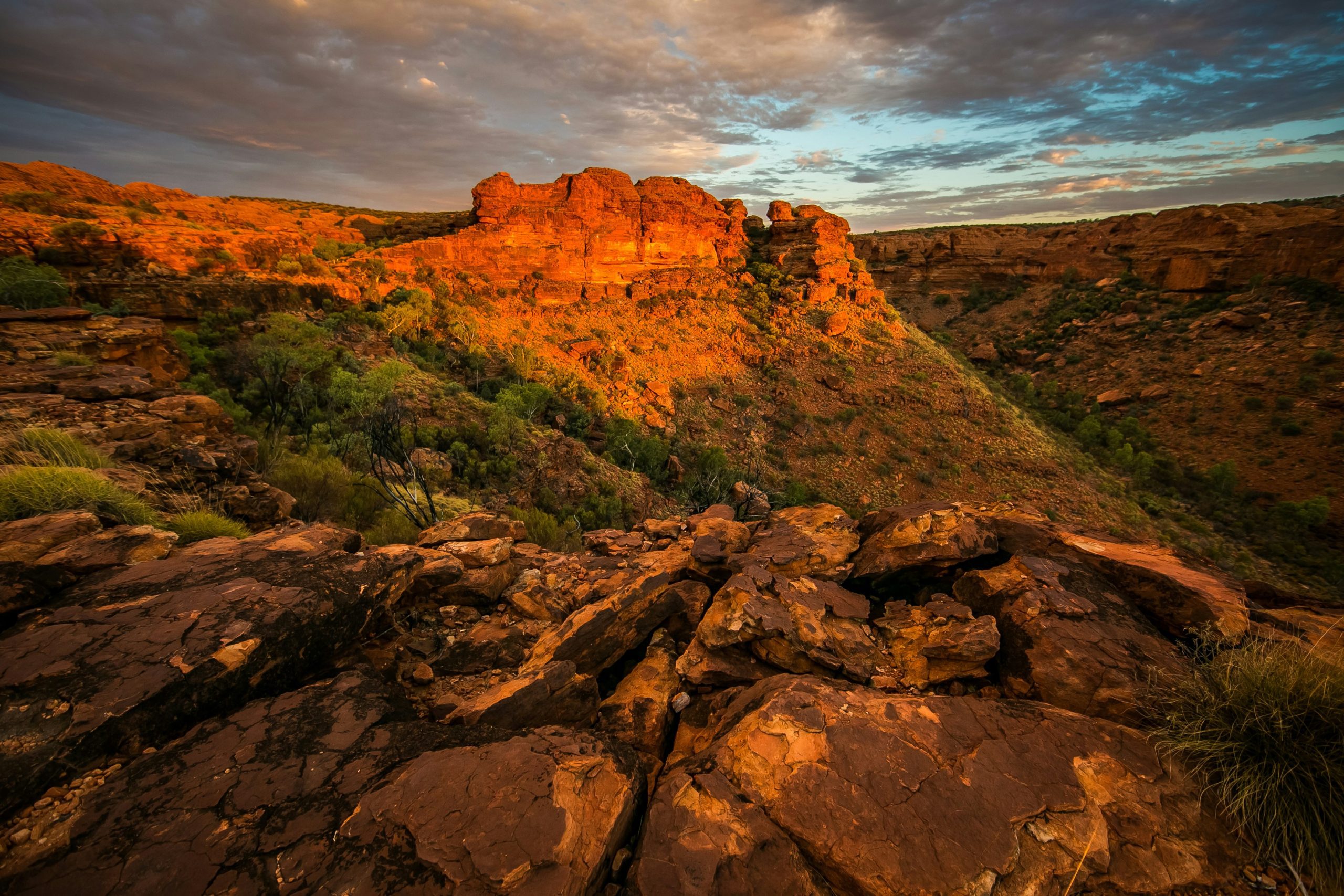 Golden sunlight on red rock formations at Australia's Kings Canyon, highlighting the rugged landscape beneath a dramatic sky.
