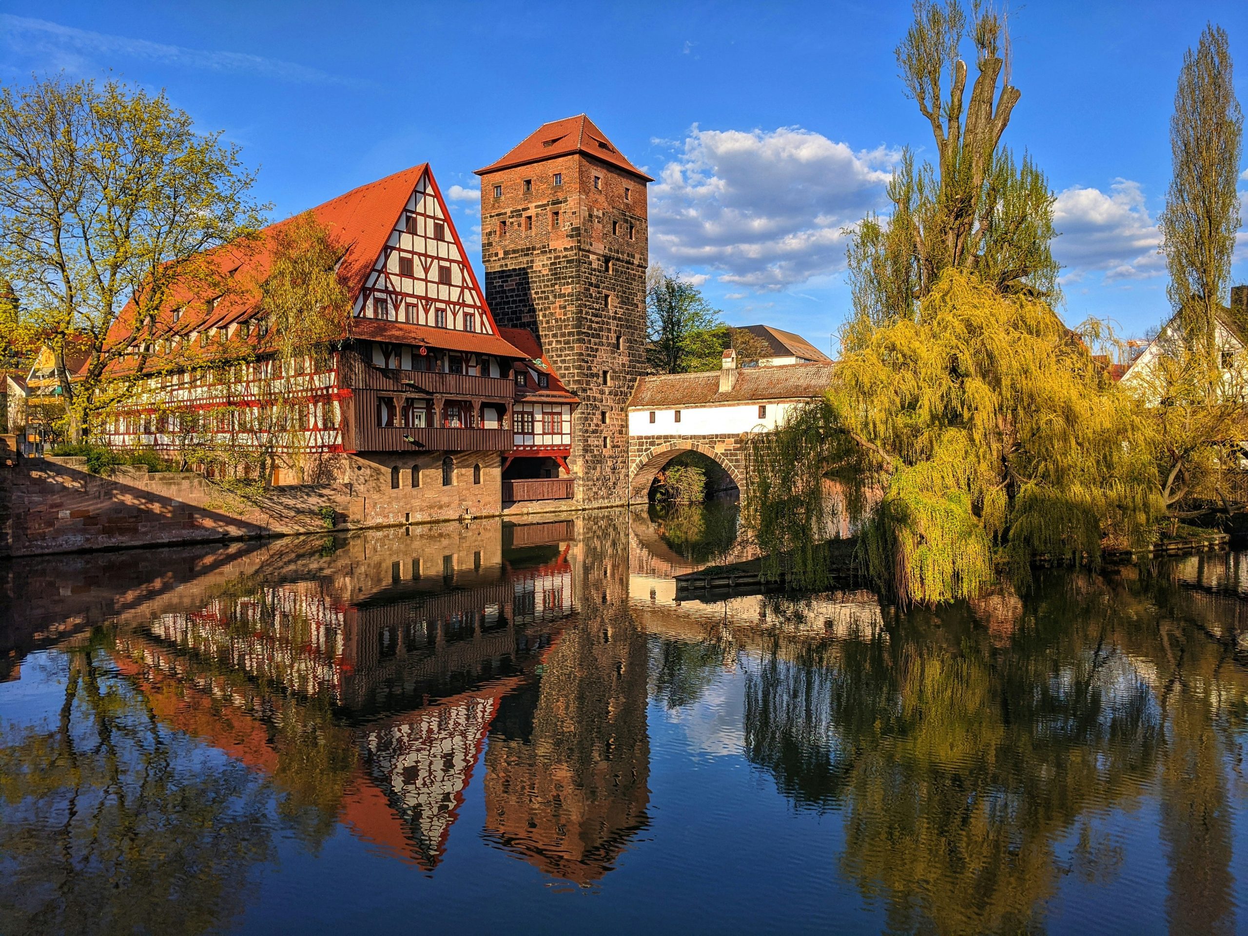 Historic timber-framed building with tower reflected in the serene waters of Nuremberg, Germany, under a vibrant blue sky.