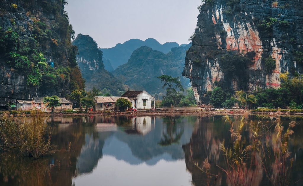 Tranquil village nestled between limestone cliffs in Ninh Binh, Vietnam, reflecting in calm waters.